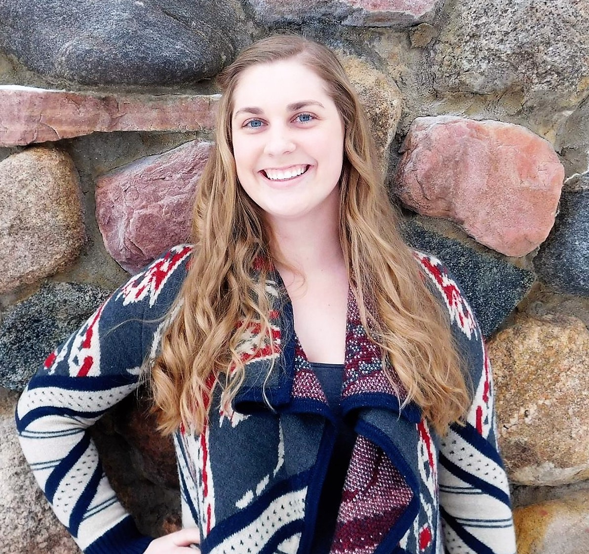 Professional photo of Madeline Schmitt, a woman with medium-length blond hair, glasses, and a smile, wearing a dark top standing in front of a stone wall.